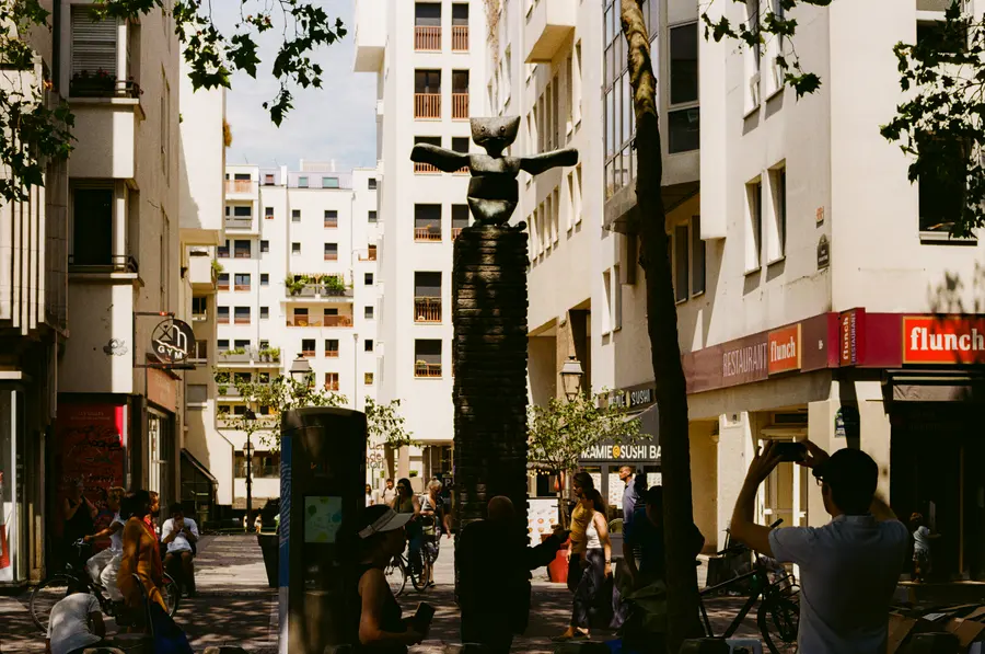 La place — Une sculpture noire au milieu d'une place ordinaire, avec les enseignes autour.