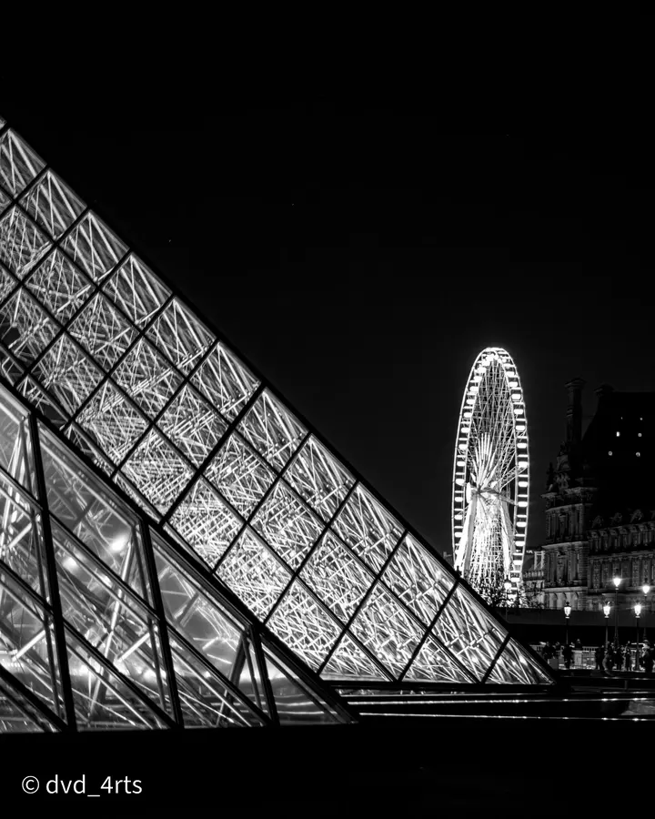La pyramide et la roue — La pyramide du Louvre découpée sur le noir, la grande roue au fond.